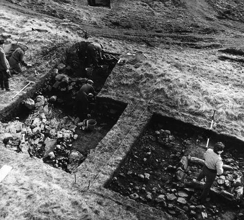 Fig 4 Hearth of the structure within the upper citadel and Rampart 1 under excavation 1959. Hearth is bottom right next to the standing excavator. Photographer was standing to the north looking south. © Historic Environment Scotland 1902341.