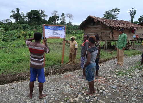 Figure 3. District Head of Mabül with sign listing budget and labour arrangements for the new lane of an outlying village, August 2017. This Papuan man from fifty miles southeast has been posted to a new District comprising the northwest third of the Korowai area. He was touring upstream villages to inspect labour projects, for reporting to Asmat Regency.