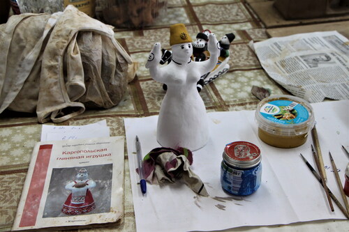 Figure 8. A work desk from the “Bereginya” craft centre: the surface is covered with oilcloth and white paper, a Bereginya toy standing in the centre, it is already fully covered in white colour paint, but only her head, face and birds have colour and pattern, the rest is not painted yet. There are thin brushes, a pen, small jars, and pieces of cloth with stains of clay laying around the toy. Photo by V. Kobyshcha.