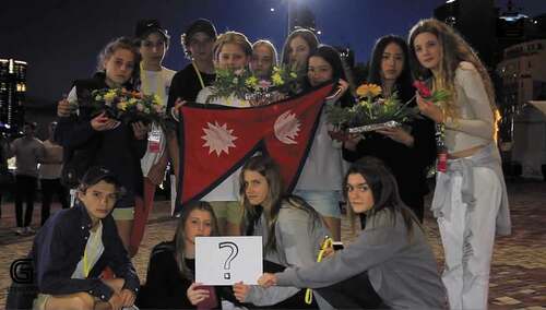 Figure 3. A group of young people in Melbourne assemble around a graphic representation of the title of the song “Kina”, holding flowers and a Nepali flag in a gesture of international solidarity.  From video of Uniq Poet’s “Kina?”