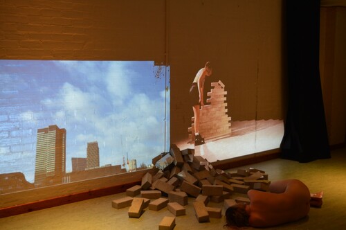 Figure 6. (Student A) shows two large projected images on a wall. Firstly, a number of bricks placed on top of each other with the performer (a white young woman wearing black shoes and shorts and a cream crop top) placing bricks on top of each other with one brick falling from the top of the tower. Secondly, a video projection overlooking London near Grenfell Tower. In front of the projections are a pile of bricks placed as though they are rubble, and the performer lies naked live in the space on her side with her back to the audience.