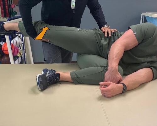 Figure 4 Gaenslen Test (modified technique) – With the patient lying in a lateral decubitus position, the painful SIJ is away from the table and contralateral leg is flexed toward the patient’s chest (similar to the traditional Gaenslen test). The Examiner stands behind the patient, who is positioned at the edge of the bed. Then stabilize the pelvis using one hand by applying a firm anterior pressure, and then extends the patient’s lower extremity at the hip ipsilateral to the painful SIJ as depicted by the arrow. Consider this modified test for those who are unable to lay supine.