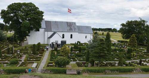 Figure 6. Emblematic Jelling as it is seen today from the top of the left gravemound, with both rune stones, encased in glass, by the church door, and a Danish flag waving behind it. The church is surrounded by family graves located in typical Danish hedged-in square parcels well-tended with greenery and flowers. Many also include natural looking, rune stone style gravestones