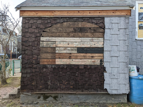 Figure 6. A view of the side vestibule after asphalt shingle siding had been partially stripped, revealing original window shapes boarded over, and original 1886 cedar shingle details. Photograph by Kris Manjapra.