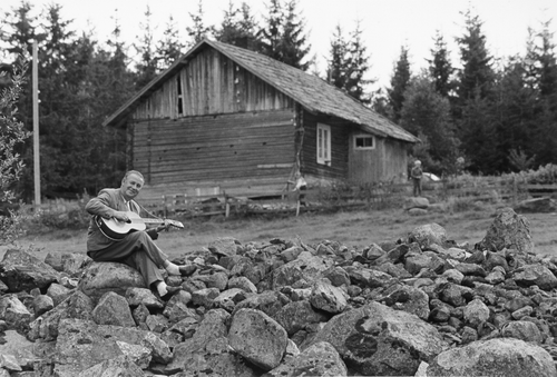 Figure 6. Alf Prøysen, as an adult, playing the guitar while sitting on a large, clearance cairn below his childhood home; Prøysenstua. Photo: Johan Brun – Domkirkeoddens fotoarkiv (dig-009776).