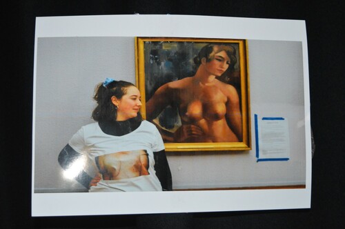 Figure 2. (Student D) shows a young white woman with her blonde hair tied back, who is wearing a white T-shirt with an image of breasts that have been through mastectomy surgery printed on it. She is posing in a way that imitates a painting behind her – the painting is of the top half of a naked woman with both breasts.