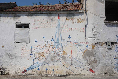 Figure 6. Graffiti in the village of Germiyan, Urla-Çeşme, protesting about environmental destruction. The village is shown at the centre; the text reads ‘We do not want exploding quarries, whizzing wind turbines, and fish farms that pollute our seas’ (top), and ‘Our villages are our places for living’ (left). (Photo and translation by Elif Koparal, August 2018)