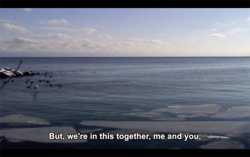 Figure 4. Still frame depicting a colour photograph of a calm body of water from the rocky shore to the horizon, under a blue sky with a few clouds. A caption reads ‘But we’re in this together, me and you’, from Me & You by Kirsty Liddiard.