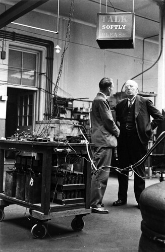 Figure 6. This famous photograph of Rutherford and Ratcliffe of 1932 was taken by Wynn-Williams who switched on the illuminated sign ‘Talk Softly Please’ just before Rutherford's arrival. The trolley contains the rebuilt Wynn-Williams and Ward amplifier as well as the three power supply batteries, enabling them to be moved from one laboratory to another (Lewis Citation1984).