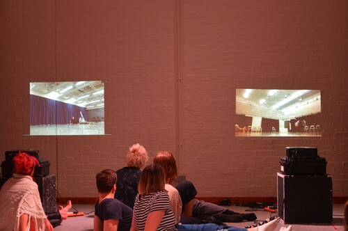 Figure 4. (Student C) shows two projected images on a white brick wall in a studio with the backs of the audience visible in the foreground. The projected images are moments from video projections of various people moving in a studio dance workshop.