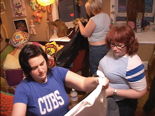 Image 7. Allyson is holding up a shirt and looking at it. Joanne is looking at it with her. Zoe is putting an item from the swap in a bag.