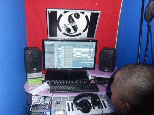 Figure 1 Studio of Seres Produções run by DJ Satelite, Luanda, 2012—Studio of Seres Produções run by DJ Satelite in a storeroom in a residential building. On the bottom screen a work-in-progress afrohouse track in FL Studio software. Top screen shows the produtora’s logo. The set-up comprises desktop PC, a two octave keyboard as MIDI controller, two monitor speakers, external audio interface, two screens, an effect rack (under bottom screen), headphones, five channel mixing board (left of keyboard), and microphone with pop filter (far right). Marçal, Luanda, 2012. Photo by the author.