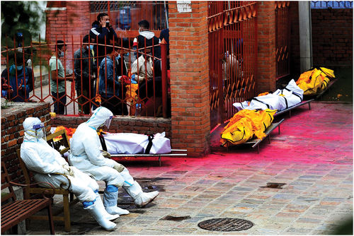 Figure 6. Members of the Nepal Army wearing PPE take a short rest while mourners performing cremation rituals outside the closed main gate of electric crematorium. Notice traces of ritual offerings with auspicious red vermillion powder on the ground. Photo by Kiran Panday®, 5th of May 2021.
