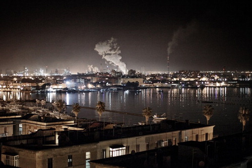 Figure 2. Night view of the Mar Piccolo area. Note the poles for mussel aquaculture in the Mar Piccolo and, in the background, the chimneys, with industrial discharge, of the ILVA steel plant and the ENI oil refinery. Photo courtesy of Mattia Insolera.