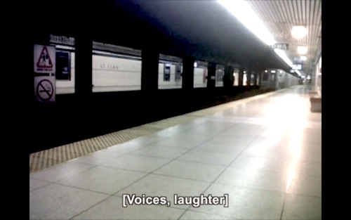 Figure 5. A still frame showing an empty subway station platform as a train leaves the station, with a caption reading ‘[Voices, laughter]’ appearing at the bottom of the frame. From Leaving Eustachian by jes sachse.