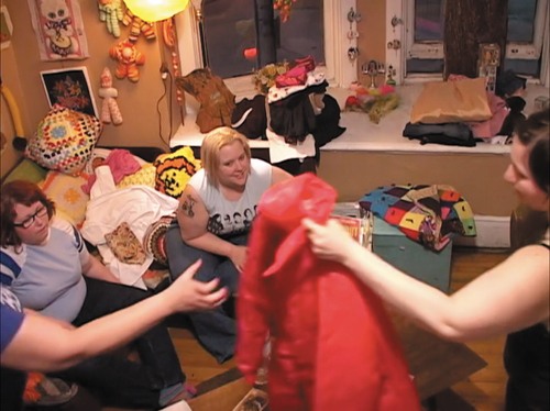 Image 2. Allyson’s hand extends to grasp a red jacket as Tracy reaches out to pass the garment to her. Joanne and Zoe sit on the couch, observing the exchange and conversingFootnote3.