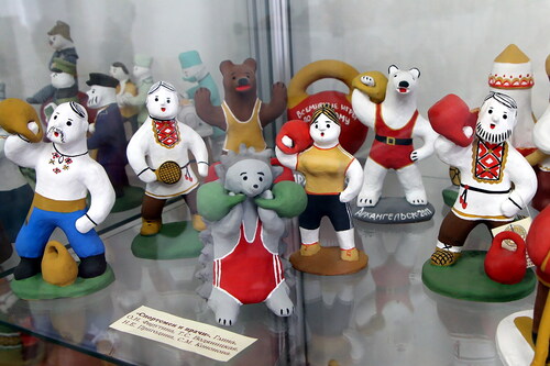 Figure 10. A glass shelf at the Centre for Folk Crafts “Bereginya” with several bespoke clay toys representing various sport activities. Men in traditional peasant outfits, bears and A glass shelf at the Centre for Folk Crafts “Bereginya” with several bespoke clay photograph, stands a female athlete who holds a kettlebell, has a stocky body, is dressed in short-sleeve top and shorts, and stays steadily on two separate legs. Photo by V. Kobyshcha.