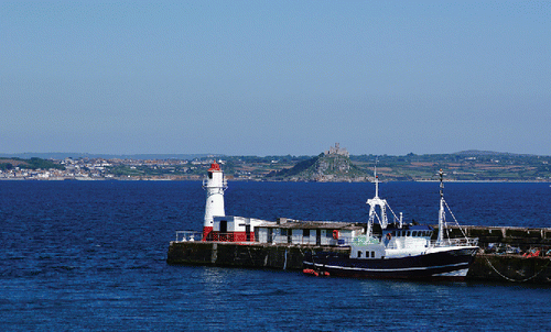 Figure 2. A view to the north-east, from Newlyn Harbour to St. Michael's Mount, showing the lighthouse and Tidal Observatory to its right, both painted red and white. The white GPS antenna on top of the lighthouse platform can be clearly seen. Photograph taken in 2010. A Wikimedia Commons image.