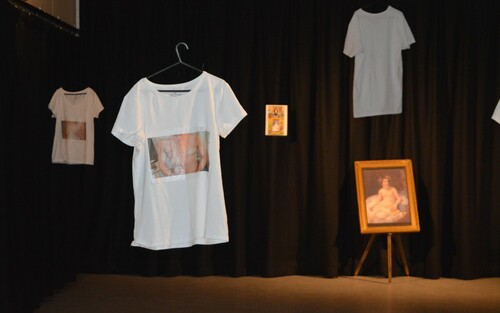 Figure 1. (Student D) is an image of a performance installation, containing white T-shirts with images of breasts that have been through mastectomy surgeries printed on them. There is a painting propped up on an easel of a naked woman in the background. There are dark curtains at the back.